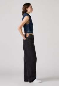 Gilet en denim bleu foncé, avec un col et sans manches ; associé à un pantalon large noir taille haute ; baskets blanches.