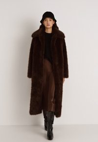 Hnedý dlhý kabát z umelého kožúšku so širokým golierom, skombinovaný s čiernym topom, hnedou sukňou a čiernymi čižmami po kolená. Modelka má na hlave čierny klobúk.