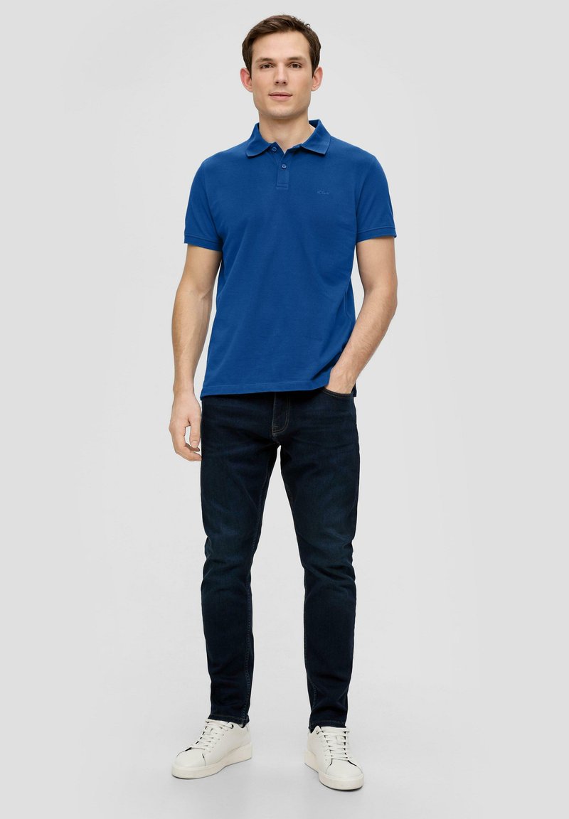 Polo blu in cotone, abbinato a jeans blu scuro e sneakers bianche. Design semplice con colletto e maniche corte.