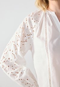White blouse with embroidered, broderie anglaise long sleeves. Features a vertical decorative trim and soft, textured fabric.