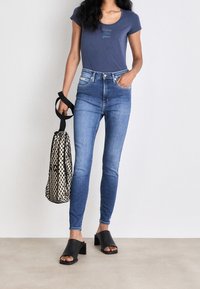 Camiseta de algodón azul oscuro con cuello redondo, pantalones ajustados de mezclilla azul claro, sandalias negras de tacón bloque y una bolsa de malla negra.