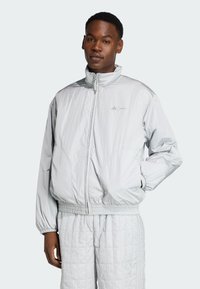 Chubasquero gris claro con cuello alto, puños elásticos y dobladillo. Material suave y brillante con un sutil logotipo en el pecho. Combinado con pantalones a juego.