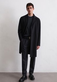 Mann in schwarzem Überrock, dunklem gerippten Pullover, schwarzen Hosen und schwarzen Lederschuhen, der an einer weißen strukturierten Wand steht.