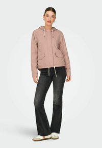 Femme portant une veste à capuche rose poussiéreux, un jean noir évasé et des baskets blanches, debout dans une posture détendue sur un fond uni.