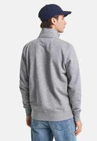 Sudadera gris con cuello alto y mangas ajustadas. Textura suave, corte relajado. Combinada con jeans de mezclilla azul y una gorra azul marino.