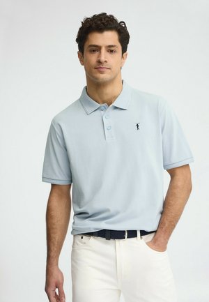 Uomo con capelli ricci scuri che indossa una polo azzurro chiaro con piccolo logo ricamato e pantaloni bianchi, mano in tasca, sfondo neutro.