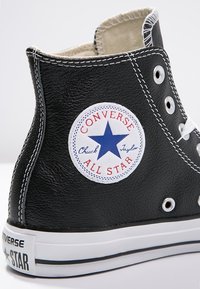 Czarne skórzane wysokie tenisówki Converse, ukazujące białą gumową podeszwę, metalowe oczka oraz okrągłą naszywkę z logo "Converse All Star Chuck Taylor".