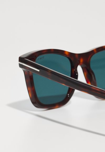 Lunettes de soleil écaille avec des verres bleu foncé, montrant un gros plan de la face avant et d'une branche sur une surface blanche.