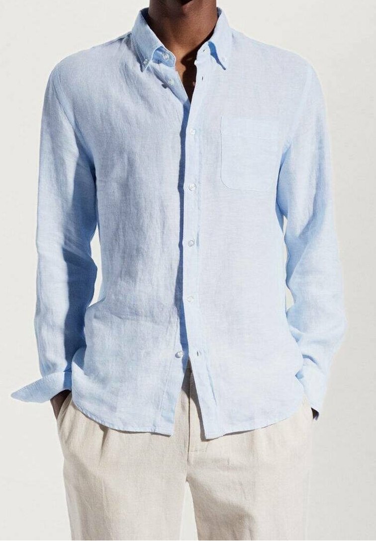 Camisa de lino azul claro con cuello abotonado, mangas largas, un bolsillo en el pecho y un corte relajado, combinada con pantalones de color claro.