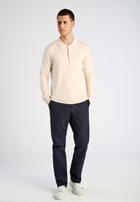 Vaalean beige pitkähihainen poolopaita, jossa on kaulus, yhdistettynä tummansinisiin housuihin ja valkoisiin lenkkareihin. Poolopaidassa on hienovarainen kiilto ja pieni logo.