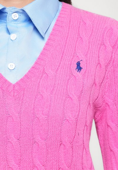 Roza pletena jopica z V-izrezom in rebrastimi detajli. Na levi strani prsnega koša je majhna temno modra oznaka.