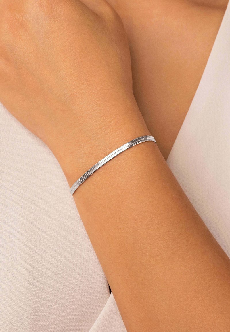 Pulsera de metal plateado con un diseño elegante y plano que presenta un centro texturizado. Lleva puesta en una muñeca sobre un fondo de tela de color claro.