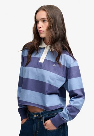 Junge Frau, die ein blau- und marineblau gestreiftes Rugbyshirt mit weißem Kragen trägt, steht mit den Händen in den Taschen und schaut zur Seite.