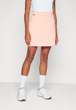 Falda deportiva de color melocotón con una textura suave, que cuenta con una cinturilla plana y bolsillos laterales, combinada con calcetines deportivos blancos y zapatillas.