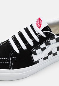 Svart semsket sneakers med et hvitt sjakkmønster på siden, hvite snørebånd og en rød Vans-logo på tungen. Glatt gummisåle.