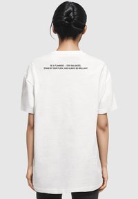 Tricou alb oversized cu maneci scurte, guler rotund și text pe spate. Materialul pare moale, cu o textură netedă.