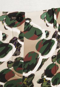 Tissu à motif camouflage présentant des nuances de vert, de marron et de beige ; comprend un détail de bouton noir et une fermeture éclair.