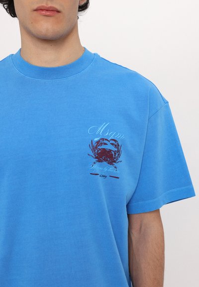 Blå bomull t-shirt med en krabbgrafik i röd och vit på vänster bröst. Designen har avslappnade kortärmar och rundad hals.