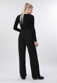 Ξανθιά γυναίκα με αλογοουρά φοράει μαύρο τοπ με μακριά μανίκια, μαύρο παντελόνι με φαρδιά πόδια και μαύρα παπούτσια, στέκεται γυρισμένη με την πλάτη σε λευκό φόντο.