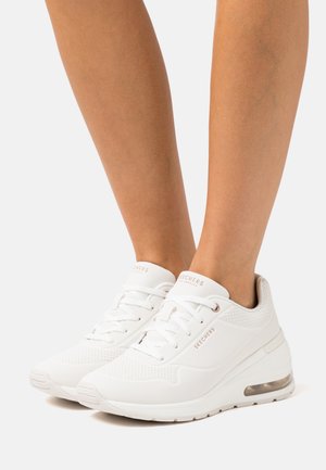 Weiße Sportschuhe mit glattem, synthetischem Obermaterial, perforierten Akzenten, Schnürsenkeln und einer sichtbaren Luftsohle in der Ferse für Dämpfung.