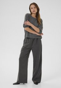 Pull gris en tricot avec des manches retroussées et des boutons décoratifs. Pantalons gris à taille haute et jambes larges. Des chaussures noires à talons pointus complètent le look.