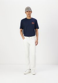 Navy T-shirt with orange graphic, white jeans, and white sneakers. Grey cap with logo, model standing against a plain white background.