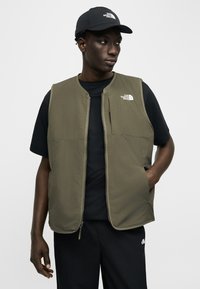 Moški, ki nosi črno kapo, črno majico, olivno zelen brezrokavnik z zadrgo in črne hlače, stoji z eno roko v žepu brezrokavnika in gleda vstran.