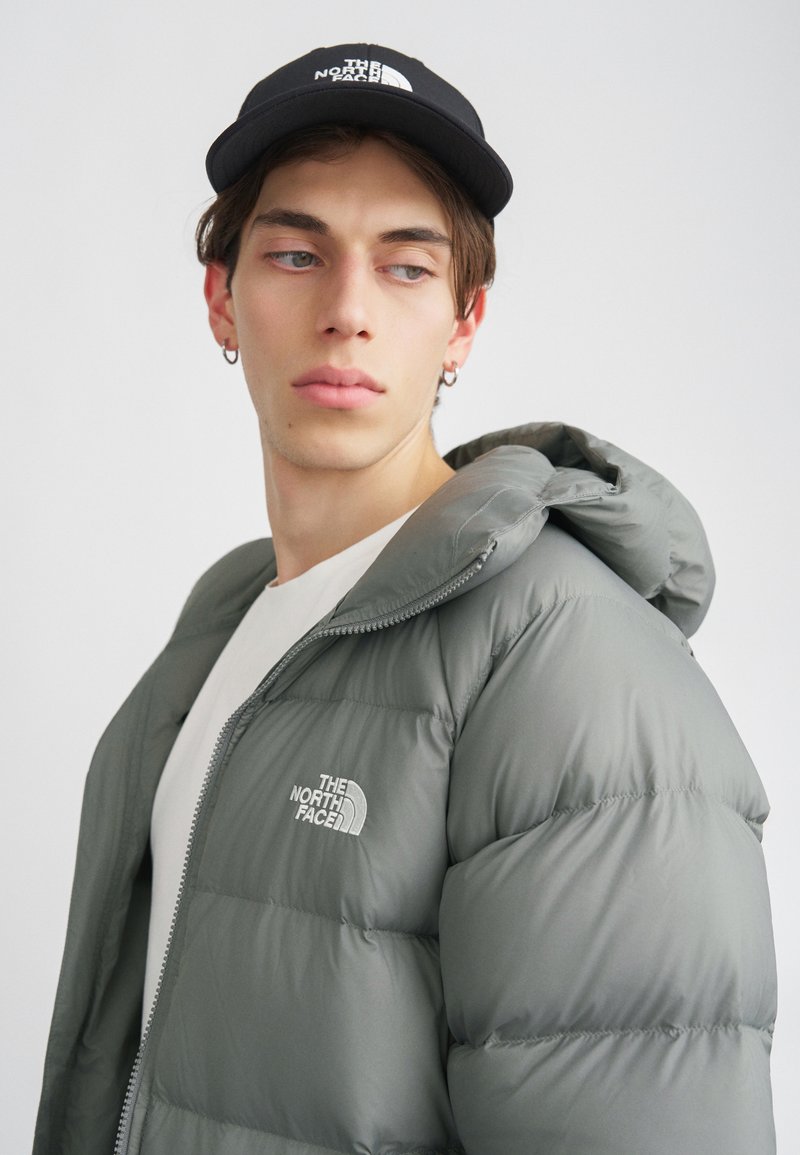 Szürke puffdzseki leválasztható kapucnival, cipzáras záródással és hímzett logóval. Fehér pólóval és fekete sapkával párosítva.