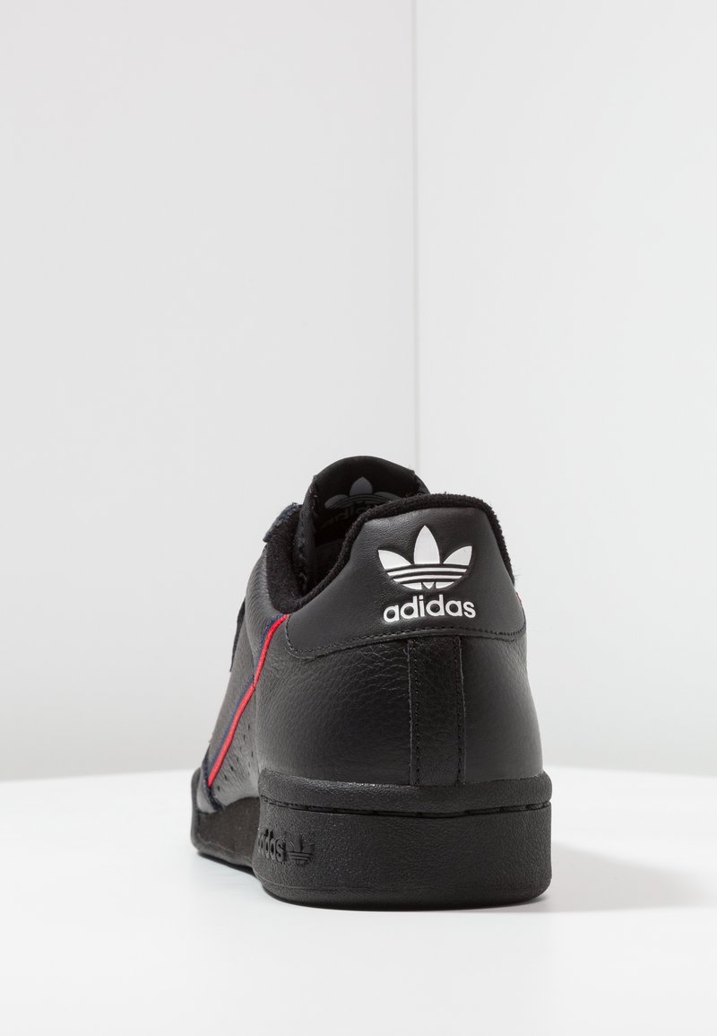 Zapatilla de cuero negra con acentos rojos, un acabado texturizado y un logotipo blanco de Adidas en el talón. Diseño redondeado con un perfil bajo.