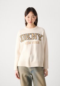 Kremfarget strikket genser med "DKNY NEW YORK" i svart og gull bokstaver. Har ribbet nederkant og brede ermer.
