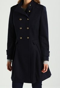 Abrigo de lana azul marino con cuello alto, diseño de doble botonadura, botones en tono dorado, bolsillos laterales y silueta estructurada. Mangas largas con puños.