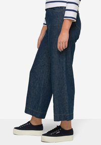 Breda byxor i blå denim med rå fåll. I kombination med svarta canvasplattformsskor. Ränder på långärmad topp syns ovanför.