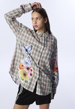 Groen en roze geruite shirt met florale en vlinderafbeeldingen. Lange mouwen met knoopsluiting, ruime pasvorm, gedragen over zwarte shorts.