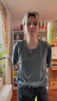 Pelēka kokvilnas T-krekls ar brīvu siluetu. Modelis stāv pretī grāmatplauktam, ģērbies melnās džinsās un ar īsiem matiem, labi apgaismotā telpā.