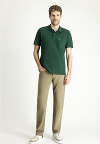 Polo verde con tasca frontale, abbinato a pantaloni beige e sneakers bianche. La maglietta presenta un logo discreto sul petto.