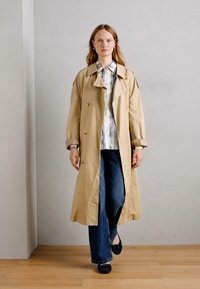 Beige trench coat, oversized szabással, dupla gombolással és nagy gallérral, csíkos inggel és sötét harangnadrággal párosítva.