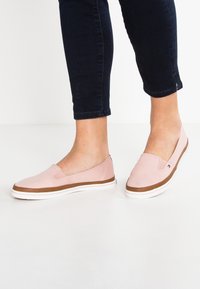 Zapatos sin cordones de color rosa claro con parte superior de lona lisa, entresuela de goma marrón y detalles de suela blanca. Diseño plano sin cordones.