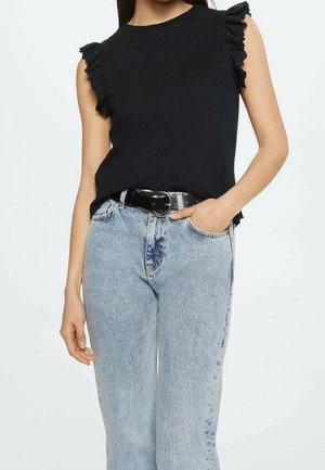 Kobieta ubrana w czarny top bez rękawów z falbankami na ramionach, włożony w jasnoniebieskie jeansy z wysokim stanem i czarnym paskiem, ręka w kieszeni.