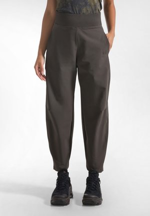 Mujer que lleva pantalones holgados de color oscuro con cintura elástica y zapatillas deportivas negras, de pie con una mano en el bolsillo.