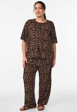 Moteris dėvi laisvą leopardų rašto marškinių tipo palaidinę su sagtukais ir derančias kelnes, stovinti ranka kišenėje ir dėvi rudus sandalų.