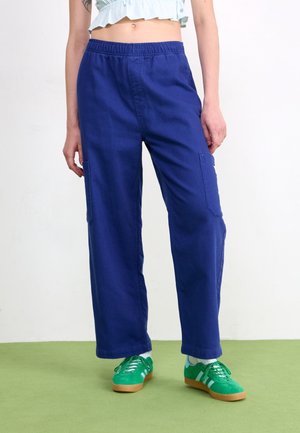 Personne portant un pantalon cargo bleu avec une taille élastique et des baskets vertes, debout sur un sol vert contre un mur blanc.