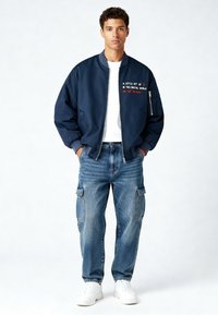 Blouson aviateur marine avec manches retroussées et texte brodé, associé à un jean cargo bleu ample et des baskets blanches.