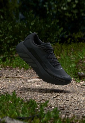 Chaussure de sport noire suspendue au-dessus d'un chemin de gravier entouré de verdure, avec de la terre et des petits cailloux soulevés en dessous.