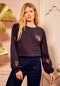 Temna dolgimi rokavi športna majica z vezeninami srca in zvezde na prsih in rokavih. V kombinaciji s temnim jeansom, ki prikazuje sproščen kroj.