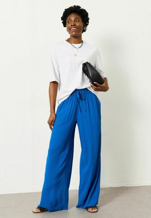 Weißes übergroßes T-Shirt, blaue weit geschnittene Hosen mit Kordelzug, schwarze Clutch-Tasche und flache Sandalen vor neutralem Hintergrund.