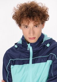 Marineblauwe en turquoise windbreaker jas met een capuchon, voorzien van een ritssluiting aan de voorkant, een mesh voering en contrasterende stikdetails.