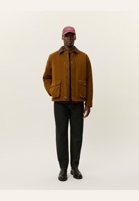 Giacca marrone con colletto marrone a contrasto, due tasche frontali, abbinata a jeans neri e stivali scuri, e un cappello bordeaux con testo.