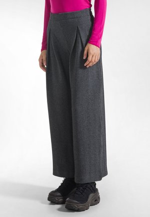 Personne portant un pantalon gris à chevrons taille haute à jambes larges, un haut rose vif à manches longues et des chaussures noires épaisses à lacets, debout sur fond blanc.
