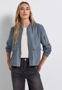 Veste légère grise avec zip à l'avant, deux poches avant et une poche à zip sur la manche. Présente des finitions côtelées et des poignets élastiques.