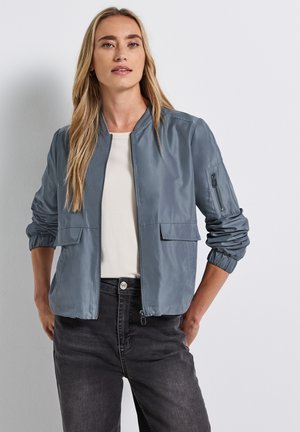 Veste légère grise avec zip à l'avant, deux poches avant et une poche à zip sur la manche. Présente des finitions côtelées et des poignets élastiques.